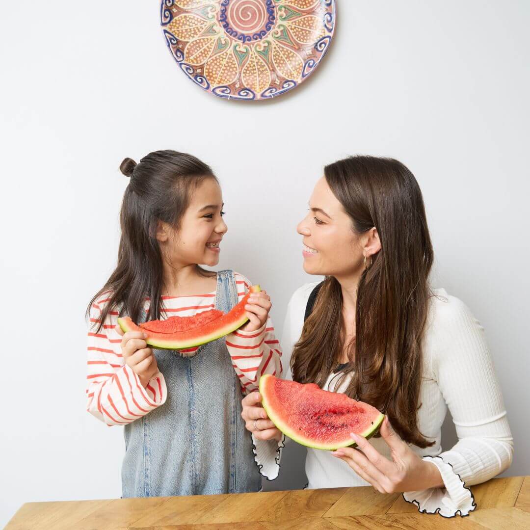 A young woman and a little girl at a table, looking at each other and smiling warmly. They are both holding watermelon. The little girl's watermelon has been eaten. They are both waring casual clothes. The woman is waaring a white tshirt with long sleves and the little girl is wearing a white long sleeve tshirt with red stripes, and a denim jumpsuit