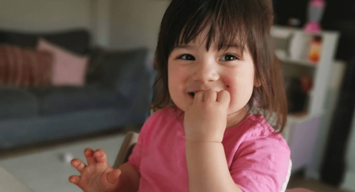 Toddler girl with dark brown hair is smiling whilst sat in a highchair eating food with her hand in her mouth and wearing a pink tshirt