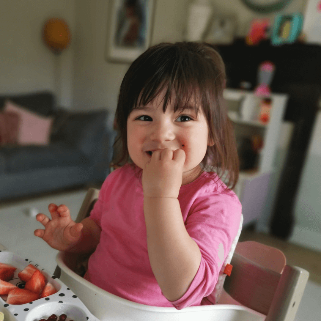 A toddler sitting on a highchair eating strawberries, wearing a pink top, looking at the camera and smiling