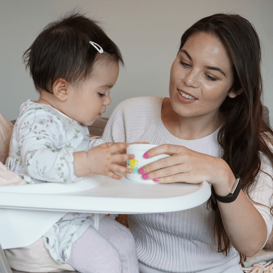 A young woman and a child sitting on a highchair. The woman is tending for the child. They look like mother and daughter. The woman is handing a sippy cup to the child.